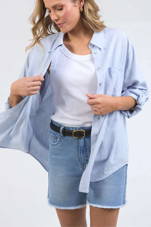 A woman with wavy blonde hair is dressed in a white tank top, an oversized light blue Foxwood Carrie Shirt crafted from breathable fabric, and blue denim shorts featuring frayed hems. Adjusting her relaxed-fit shirt with one hand, she gazes downward while showcasing a black belt accented with a gold buckle.