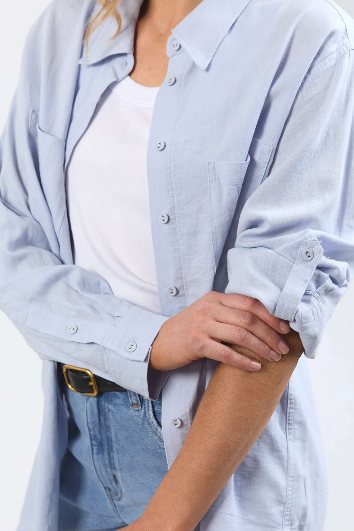 A person is shown wearing a light blue Foxwood Carrie Shirt over a white T-shirt and blue jeans. The sleeves of their shirt are rolled up, and they hold one arm with the other hand. The relaxed fit and breathable fabric of the Foxwood Carrie Shirt create a comfortable look. Only their body from the shoulders down is visible; their face is not shown.