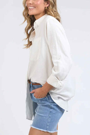 A person in a relaxed fit Foxwood Carrie Shirt and denim shorts, with one hand in their pocket, smiles against a light background.