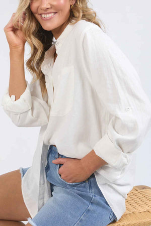 A person with wavy blonde hair smiles while wearing a Foxwood Carrie Shirt in a relaxed fit and denim shorts. They are seated on a woven chair against a plain backdrop, with the loose white linen fabric contributing to their effortlessly chic appearance.