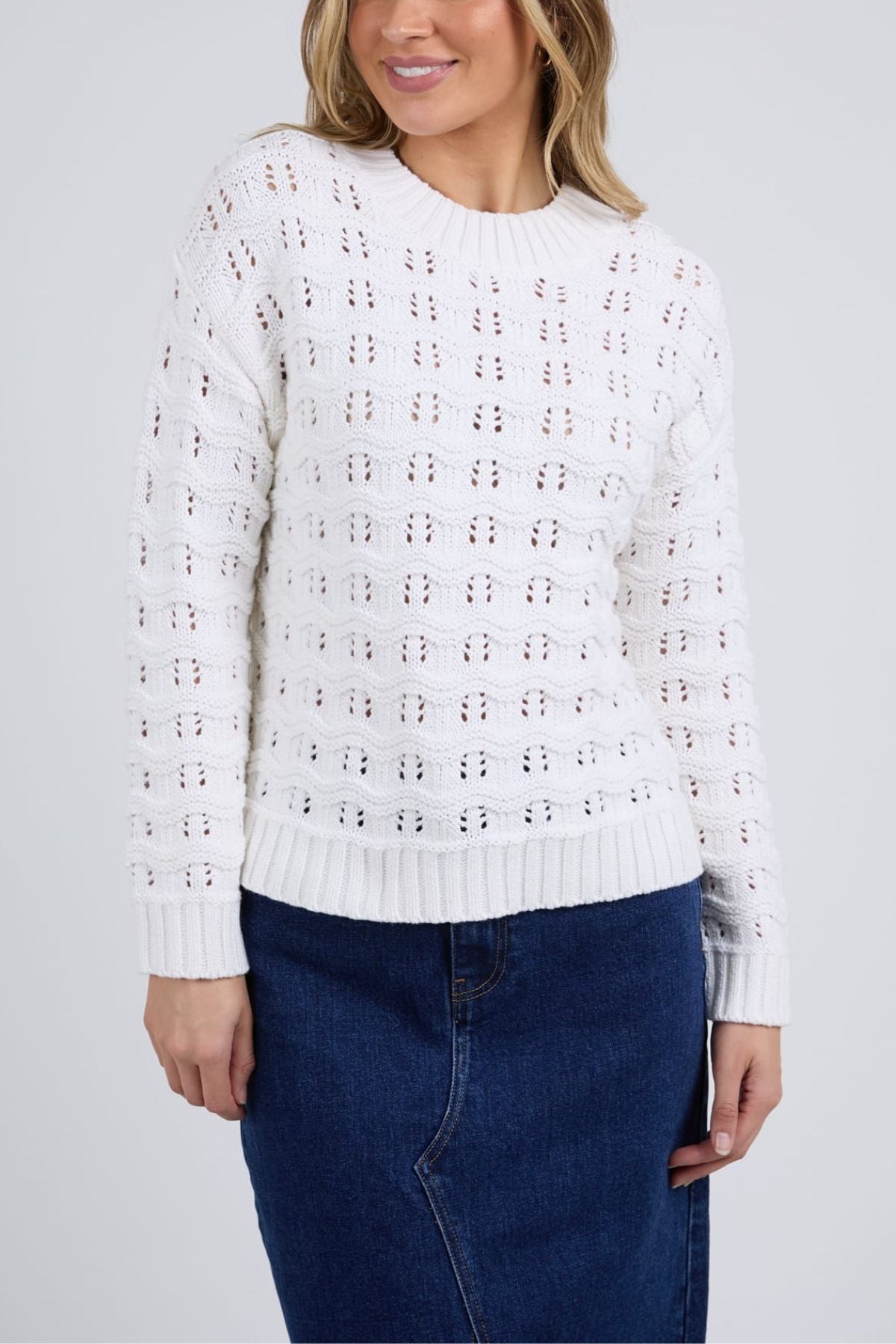A woman wears the Foxwood Carter Knit, a white long-sleeved pointelle sweater, paired with a blue denim skirt against a light gray background. Her face is partly visible and she has light brown hair—ideal for casual style.