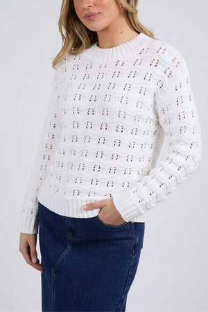 A woman stands against a plain light gray background wearing the Foxwood Carter Knit, a white pointelle sweater with a textured pattern, paired with a blue denim skirt—an ideal choice for any casual wardrobe.