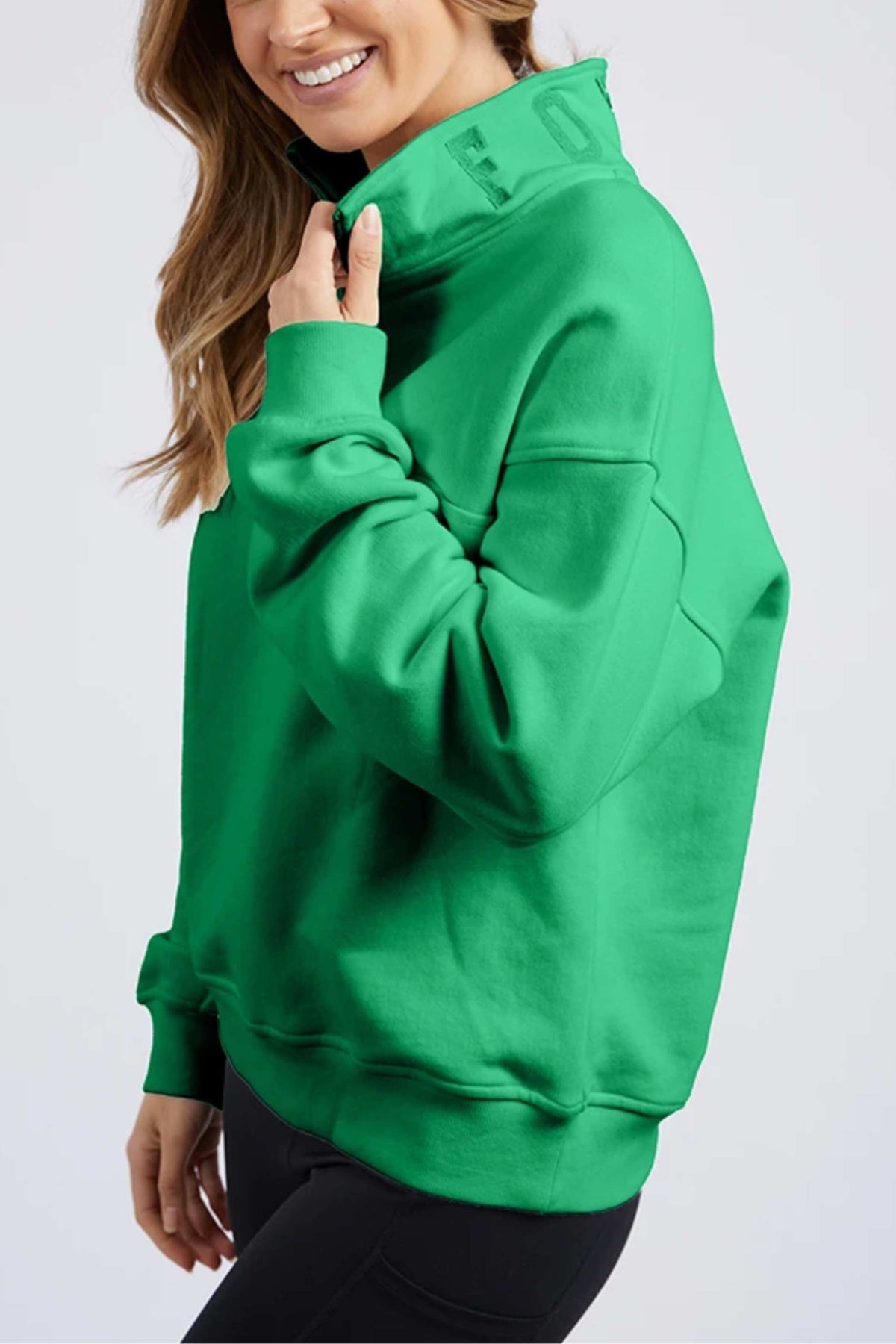 A person in a bright green Foxwood College Zip Crew with a high collar and black pants smiles while touching the collar, set against a plain light background.