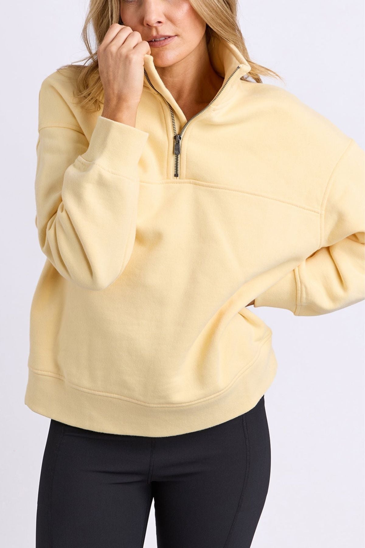 A woman wearing the Foxwood College Zip Crew in light yellow and black leggings stands against a white background, with one hand on her chin and the other in her pocket.