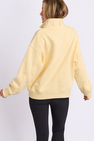 A woman with light brown hair, facing away from the camera against a plain white background, wears the Foxwood College Zip Crew in pale yellow paired with black leggings.