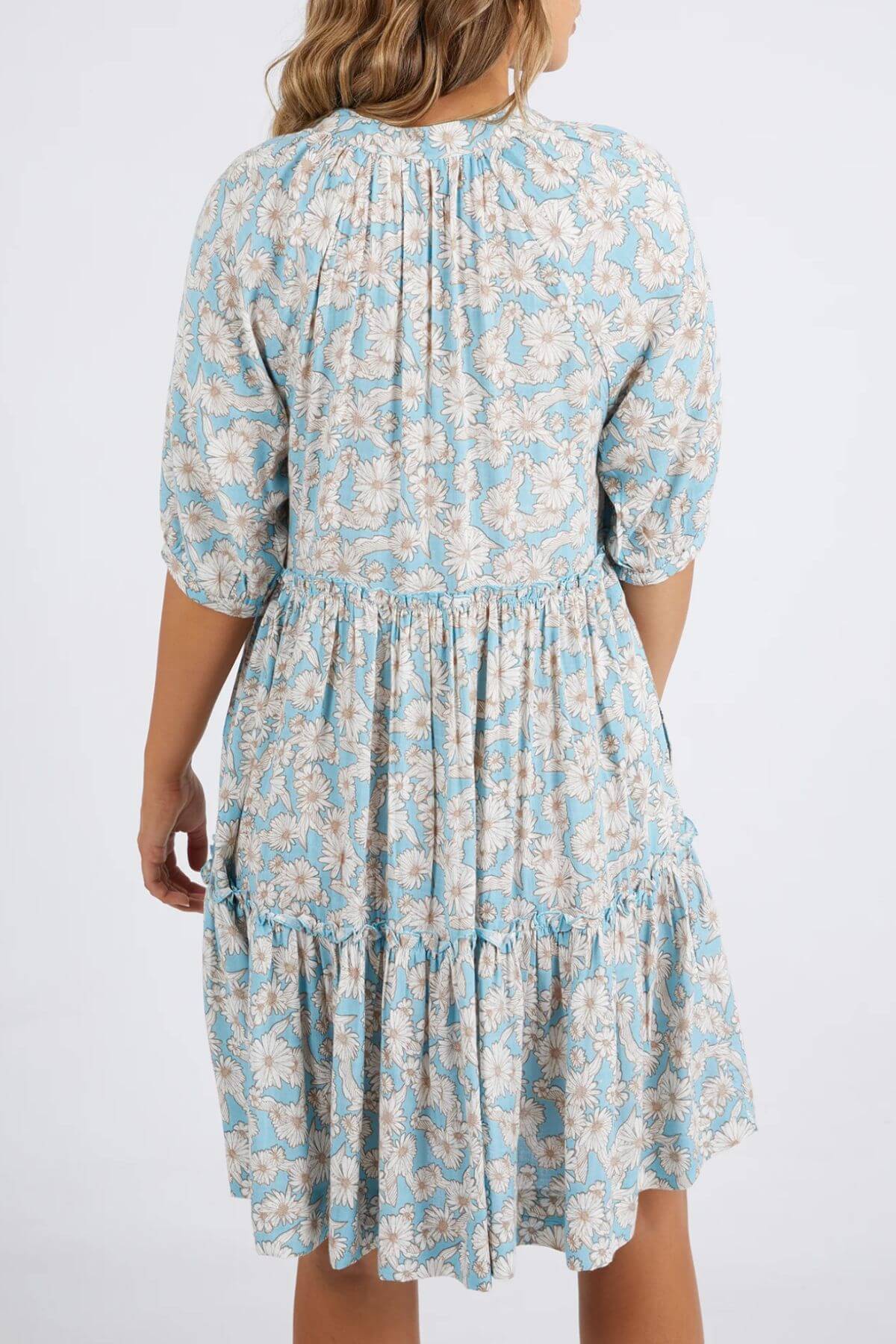 The individual is wearing a Foxwood Daisy Dress, which has a blue and white floral pattern. Made from Viscose, it features a gathered waist, elbow-length sleeves, and a tiered skirt. They stand with their back to the camera against a plain white background.