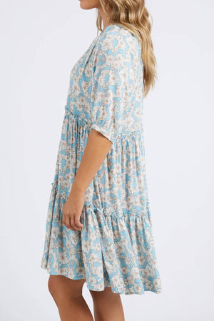 A woman with long, wavy hair stands sideways in a knee-length Foxwood Daisy Dress featuring blue and white floral patterns. The dress showcases mid-length sleeves and a tiered, gracefully flowing skirt.