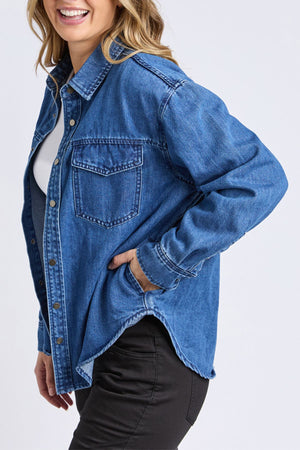 A person wears the Foxwood Daisy Overshirt in relaxed blue denim over a white top and black pants, smiling with one hand in their pocket against a plain light background.