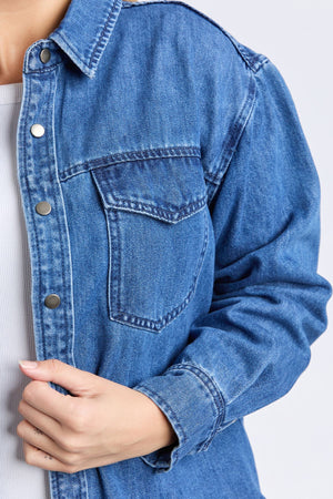 Close-up of a person in a relaxed fit Foxwood Daisy Overshirt—blue denim with metal buttons and front pocket, layered over a cotton shirt. One hand holds the overshirt near the chest.