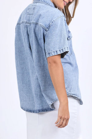 A person wearing a relaxed fit Foxwood Daisy Short Sleeve Shirt in light blue denim with a white skirt, standing sideways, head slightly turned, hair partially covering their face.
