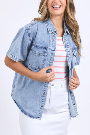 A woman smiles while wearing the Foxwood Daisy Short Sleeve Shirt, a light blue, 100% cotton relaxed fit shirt over a striped white top and white skirt, holding the open denim shirt with one hand.