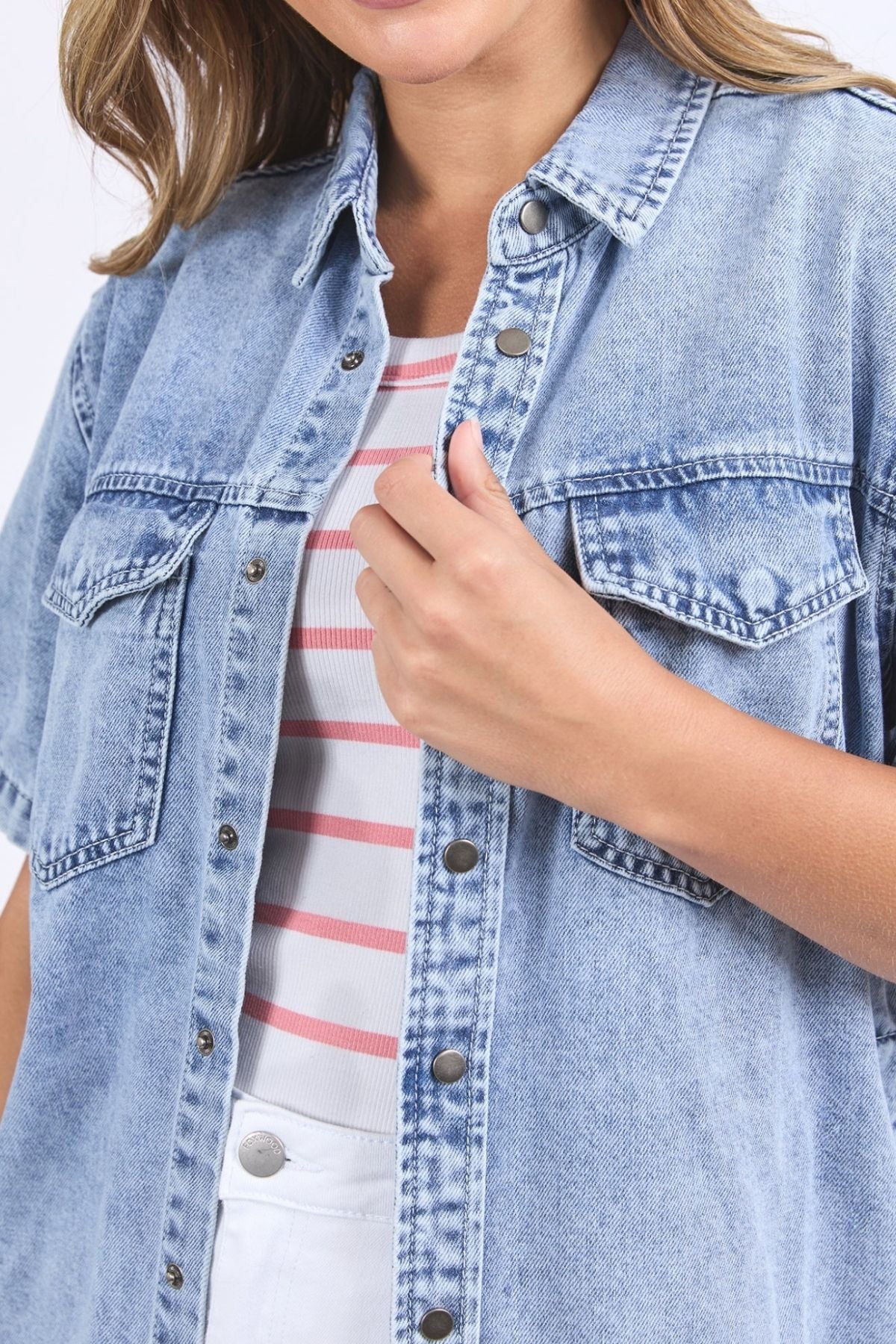 A woman wears the Foxwood Daisy Short Sleeve Shirt in light blue denim over a white and pink striped top, holding the shirt near the buttons. Her face is partially out of frame.