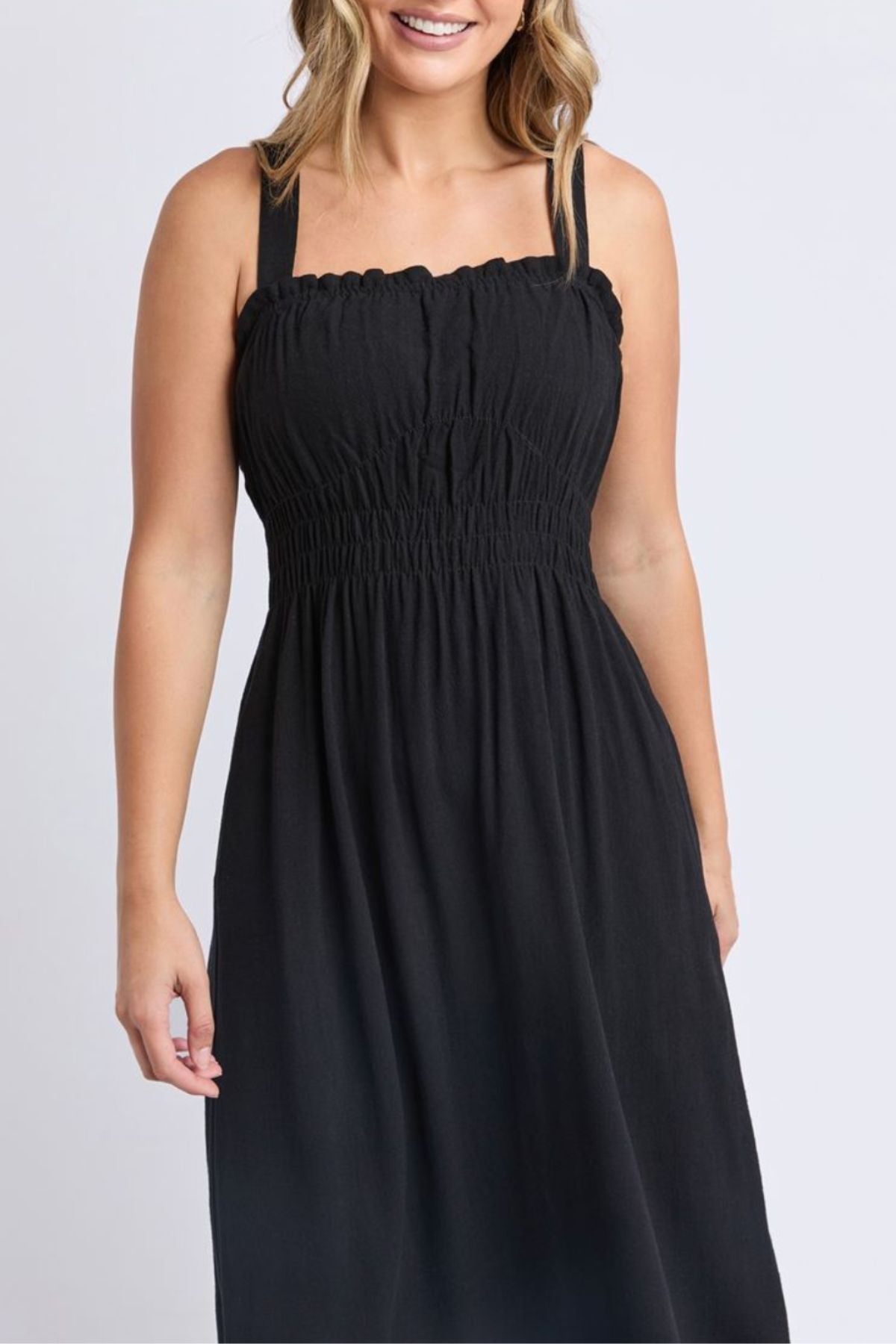 A woman smiles with her hair down, wearing the Foxwood Dandelion Dress—a sleeveless black dress featuring a smocked bodice and square neckline—while standing against a plain light background.