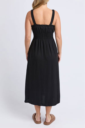 A woman faces away, wearing the Foxwood Dandelion Dress—a black sleeveless linen blend dress with wide straps and a smocked bodice—paired with tan slide sandals, set against a light plain background.