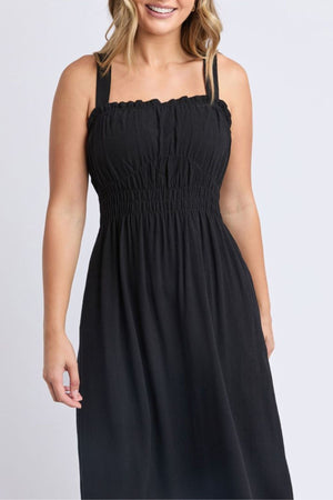 A woman smiles with her hair down, wearing the Foxwood Dandelion Dress—a sleeveless black dress featuring a smocked bodice and square neckline—while standing against a plain light background.