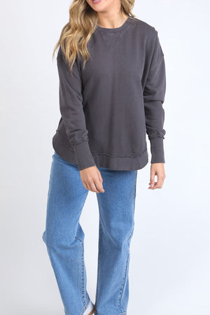 A person with long, wavy blonde hair wears a charcoal gray Foxwood Delilah Crew made from 100% cotton and light blue jeans, standing against a plain white background with their face partially hidden.