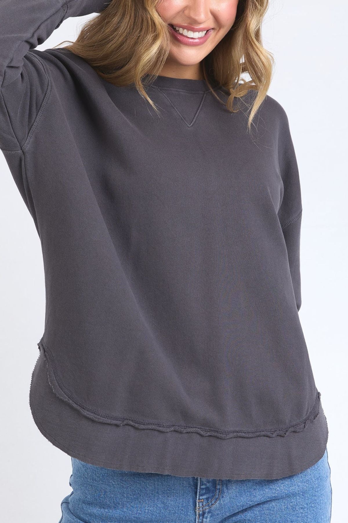 A woman smiles in a loose-fitting, long-sleeve Foxwood Delilah Crew casual jumper made from 100% cotton with raw-edge seams, paired with light blue jeans. Only her upper body appears against a plain light background.