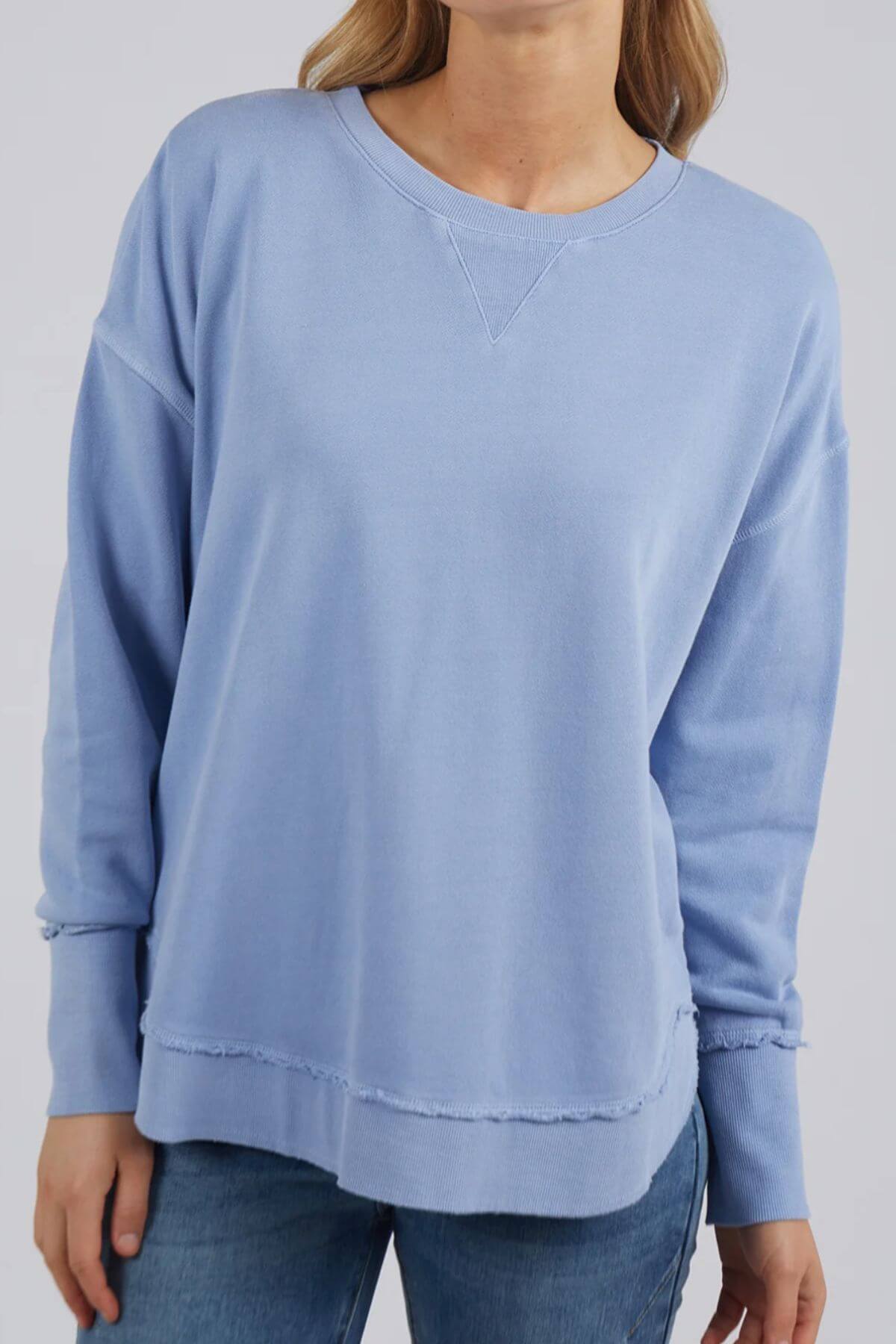 A person wearing the Foxwood Delilah Crew, a light blue, long-sleeve cotton jumper with a stitched V design and slightly frayed hem, paired with blue jeans. The plain gray background complements the outfit while the person’s head remains out of view.