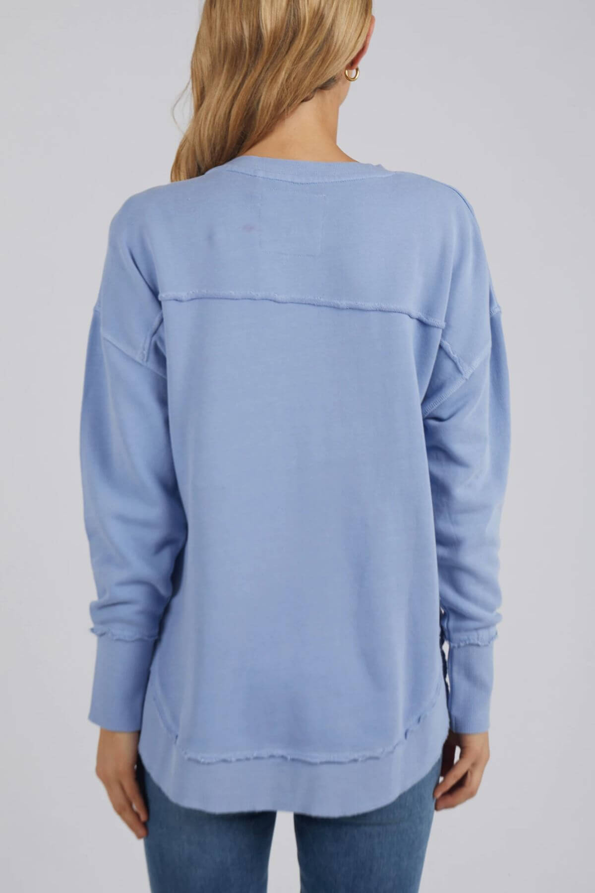 A woman with long blonde hair is wearing a light blue Foxwood Delilah Crew jumper and blue jeans, facing away from the camera.