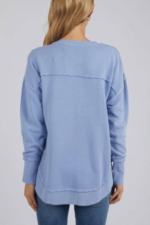 A woman with long blonde hair is wearing a light blue Foxwood Delilah Crew jumper and blue jeans, facing away from the camera.