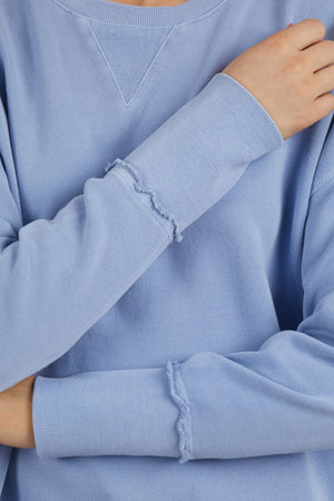 A person in a Foxwood Delilah Crew, a light blue cotton jumper with visible stitching on the sleeves, stands with arms crossed and one hand on the opposite shoulder. The neutral, out-of-focus background emphasizes the jumper's relaxed style.