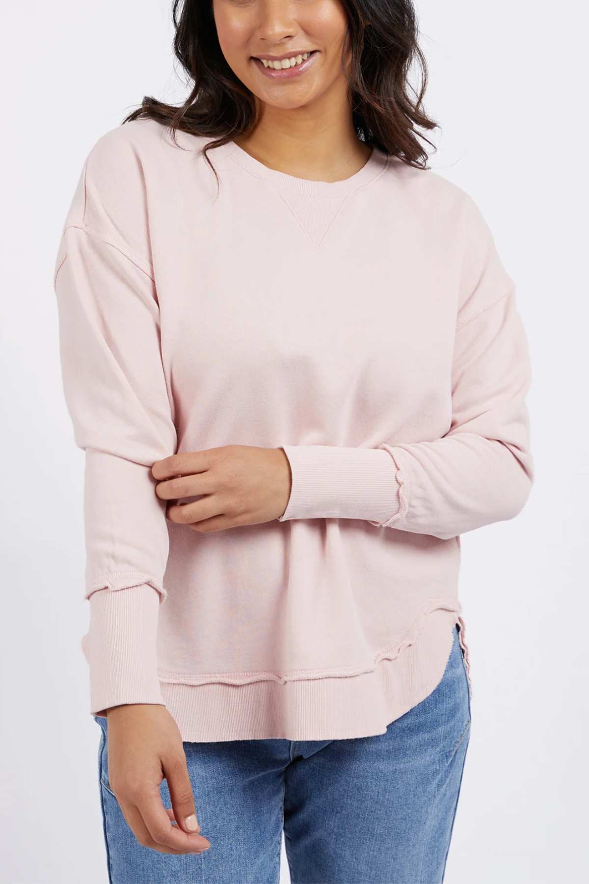 A woman wears the Foxwood Delilah Crew in light pink with blue jeans, standing against a plain background, smiling and touching her left sleeve with her right hand.