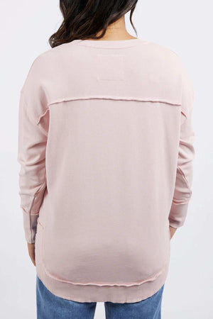 A person with long dark hair, seen from behind, is wearing a relaxed fit, light pink Foxwood Delilah Crew cotton jumper with visible seams and blue jeans.