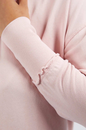 A close-up of a person wearing the Foxwood Delilah Crew, a light pink cotton long-sleeve shirt featuring a subtle wavy seam detail near the wrist. The person's hand is partially visible, touching their neck or chin.