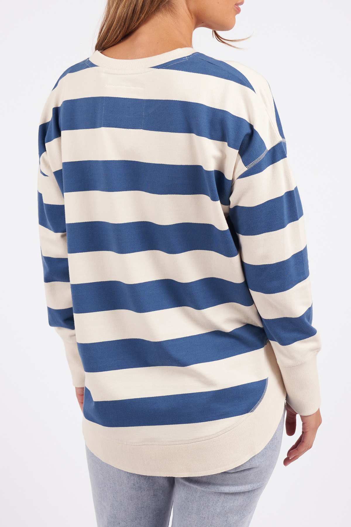 Someone wearing the Foxwood Delilah Stripe Crew, a long-sleeve cream and blue striped shirt, and light blue jeans stands with their back to the camera against a plain white background.