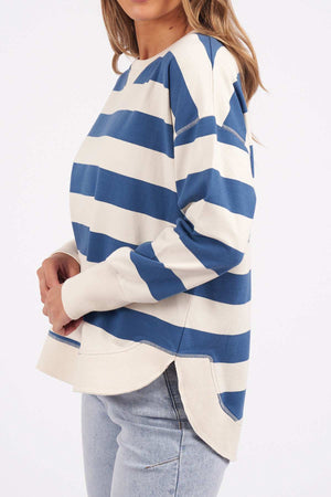 A woman stands sideways wearing the Foxwood Delilah Stripe Crew, a cotton blue and white striped jumper with a crew neck, curved side slits at the hem, and light-wash jeans. Her hair is loose.