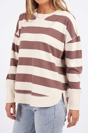 A person with long hair is wearing the Foxwood Delilah Stripe Crew, a comfortable beige and brown striped jumper with long sleeves. This 100% cotton sweater has a slightly oversized fit. The individual is also dressed in light blue jeans, against a plain white background.