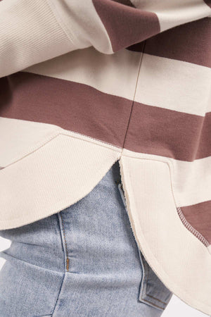 Close-up of a person wearing the Foxwood Delilah Stripe Crew, partially tucked into light blue jeans. The beige and brown striped sweater, made from 100% cotton, features a small side slit at the hem, adding a casual and relaxed look to the outfit.