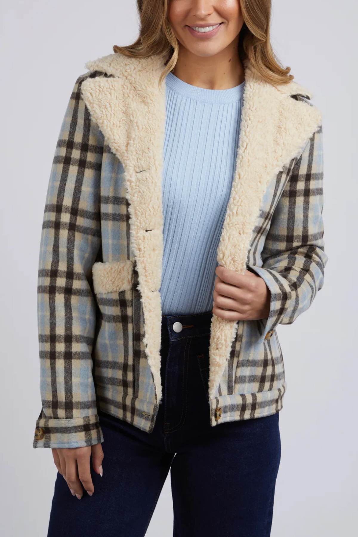 A woman wears a blue ribbed top, dark jeans, and a Foxwood Demi Check Jacket featuring light cream sherpa lining on the collar and inside, posing with one hand holding the jacket open.