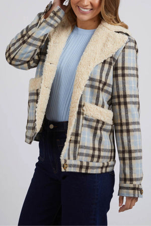 A woman in a blue sweater and dark jeans smiles while modeling the Foxwood Demi Check Jacket, featuring cream sherpa lining on the collar, lapels, and pockets, set against a neutral background.