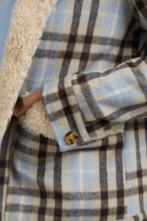 A close-up of a person wearing the Foxwood Demi Check Jacket in blue, beige, and brown checks with a fluffy cream Sherpa lining. One hand is partially tucked into the jacket, showing a brown button on the sleeve.