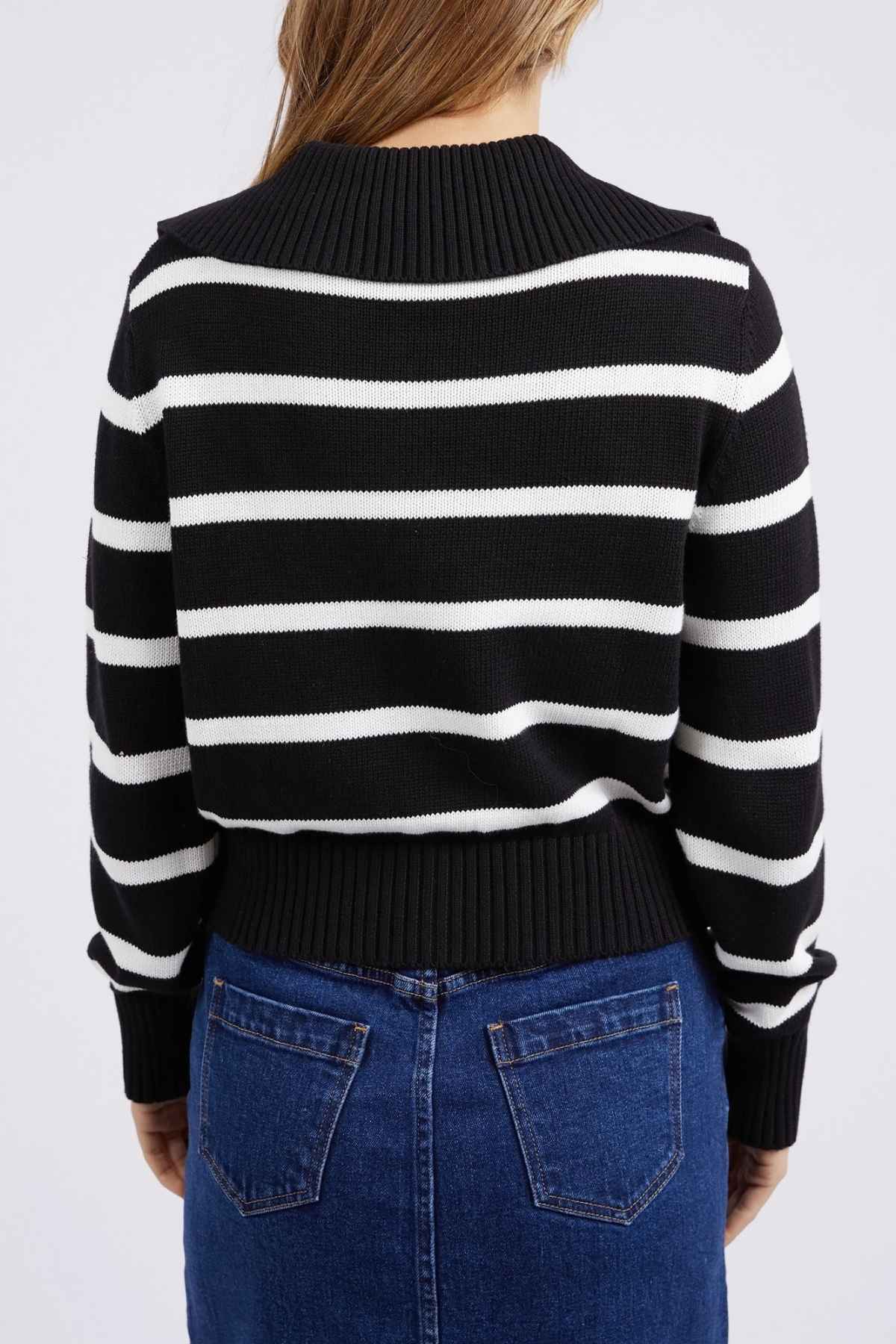 A person with long hair is standing with their back to the camera, wearing a Foxwood Effie Polo Knit sweater in black and white, paired with a dark blue denim skirt. The 100% cotton sweater boasts yarn-dyed stripes and a ribbed pattern on the cuffs and hem. The background is plain white.
