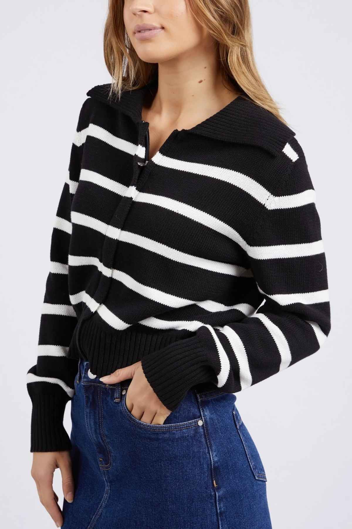 A woman wearing the Foxwood Effie Polo Knit sweater featuring yarn-dyed black and white stripes and a zip-up collar, paired with high-waisted blue jeans. She has long, wavy hair and is posing against a plain background.