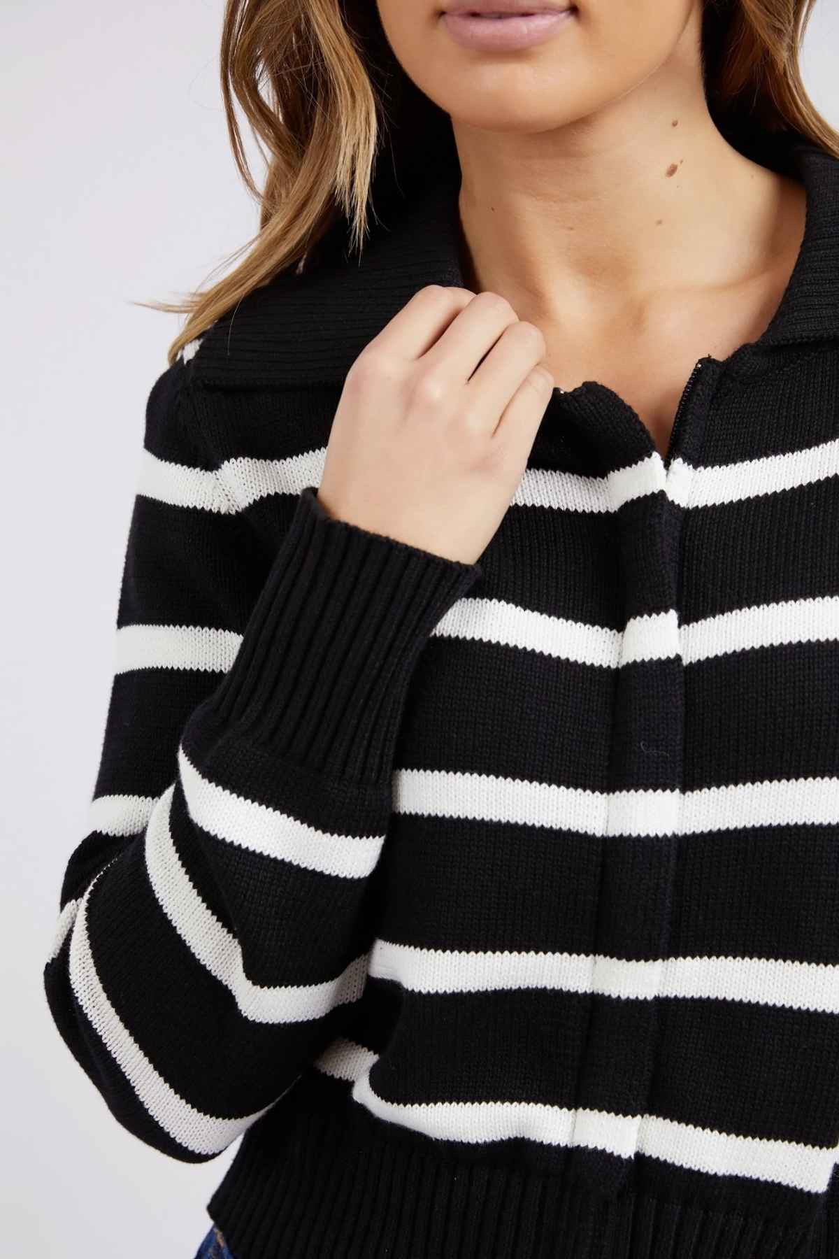 A person is seen from the shoulders to the chin, wearing a Foxwood Effie Polo Knit with black and white yarn-dyed stripes. They are holding the collar of the sweater, which is made from 100% cotton, with their right hand. The background is plain and light-colored.