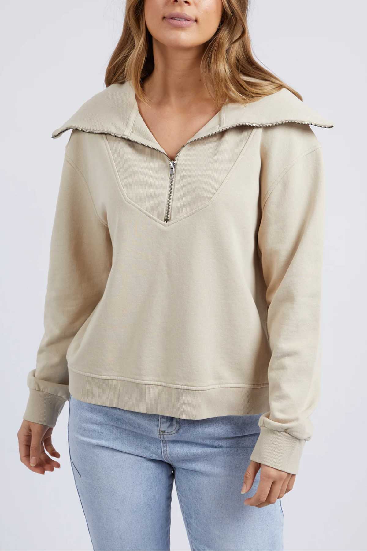 A woman wears the Foxwood Ellidy Zip Crew, a relaxed fit beige sweater in unbrushed cotton fleece, paired with light blue jeans. She stands before a plain light background with her face partly out of frame.