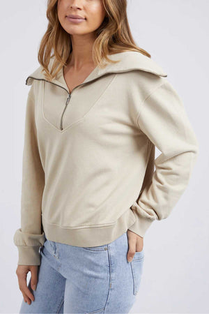 Wearing the Foxwood Ellidy Zip Crew in light beige unbrushed cotton fleece and light blue jeans, a person stands with one hand in their pocket against a plain background. The zip crew’s zipper is partly undone for a relaxed look.