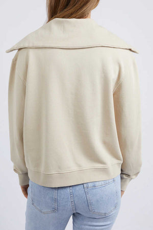 A person is seen from behind wearing the Foxwood Ellidy Zip Crew, a relaxed fit light beige cotton fleece sweater with a wide collar, styled with light blue jeans and set against a plain light background.