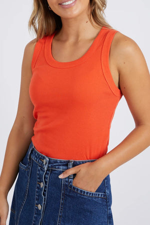 Woman wearing the Foxwood Esme Tank, a stylish orange rib knit sleeveless top, paired with a high-waisted blue denim button skirt, posing with one hand in her pocket against a plain background.