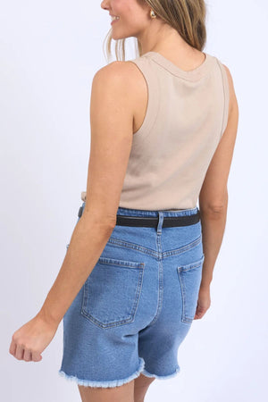A person is standing with their back turned, wearing the Foxwood Esme Tank, a beige sleeveless top ideal for summer attire, paired with blue denim shorts that have a frayed hem. The casual appearance is accentuated by the lightweight cotton blend of the tank and a plain white backdrop.
