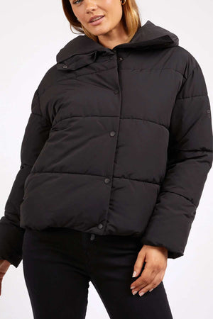 A woman models the Foxwood Essential Puffer Jacket in black with a hood and black jeans, standing against a plain white background—an ideal staple for your winter wardrobe.