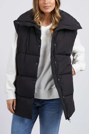 A woman in a Foxwood Essential Puffer Vest, crafted from durable polyester, wears it over a white sweatshirt and blue jeans. The vest has a high collar and snap buttons. Her long wavy hair complements the light-colored background as her hands rest partially visible at her sides.