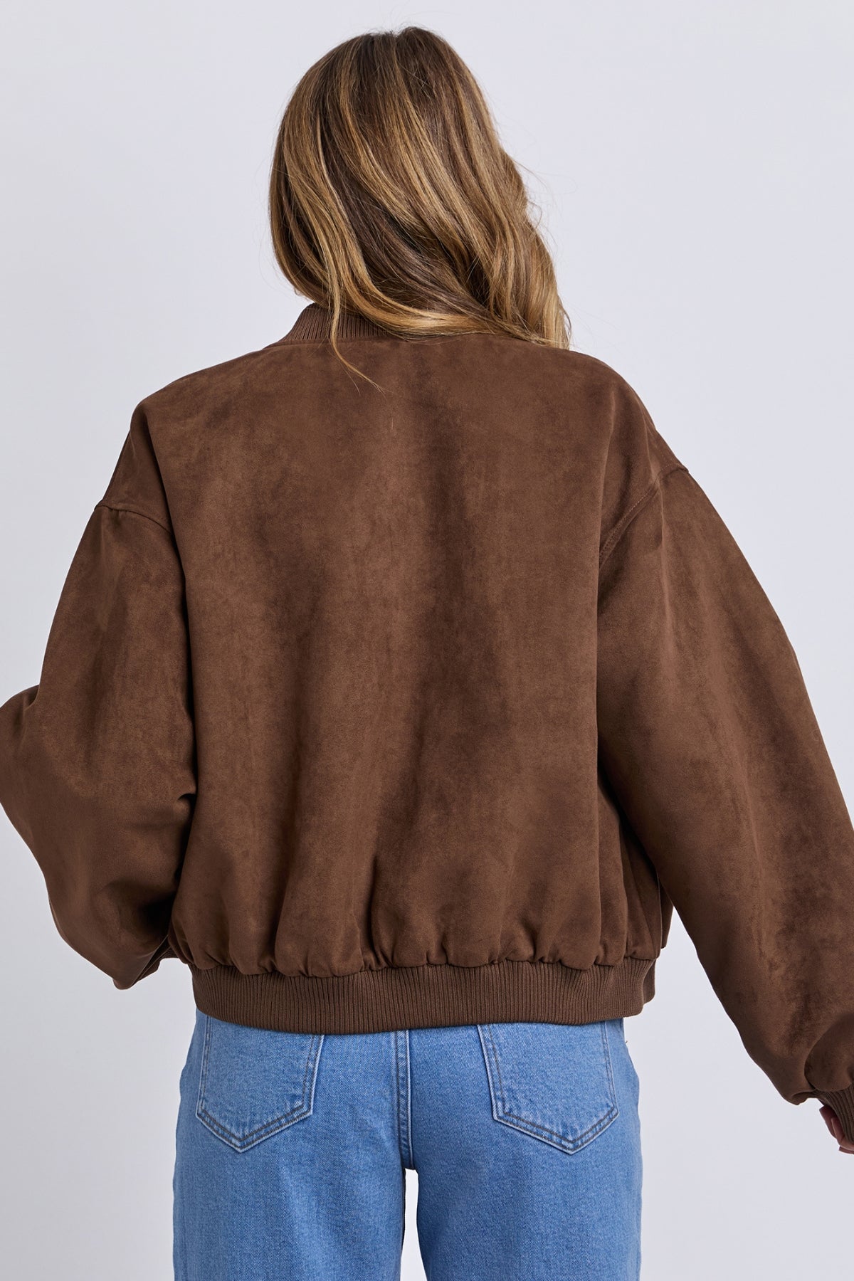 A person with long, wavy hair is seen from behind wearing the Foxwood Evie Suede Bomber in brown and blue jeans, standing against a plain, light background.