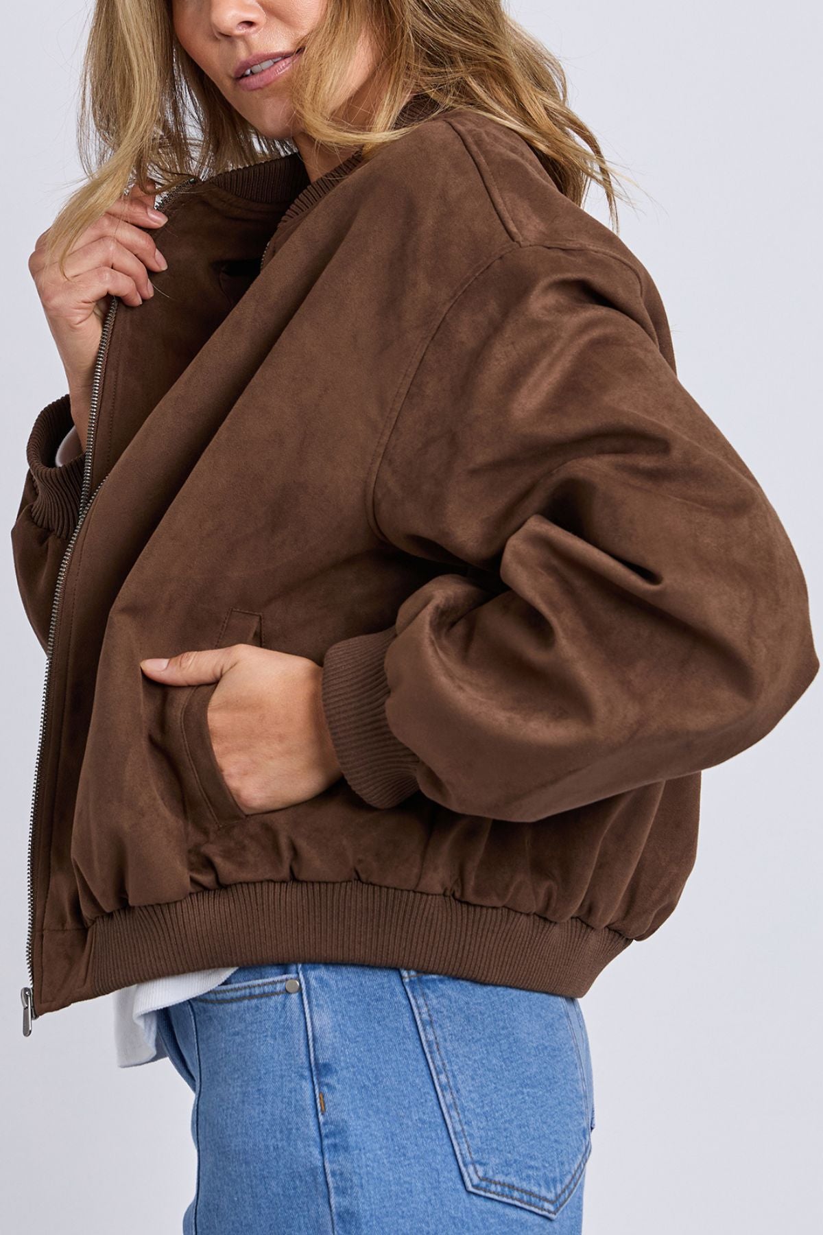 Woman wearing the Foxwood Evie Suede Bomber with ribbed cuffs and hem, blue jeans, and a white shirt, holding the jacket collar while standing against a plain light background.