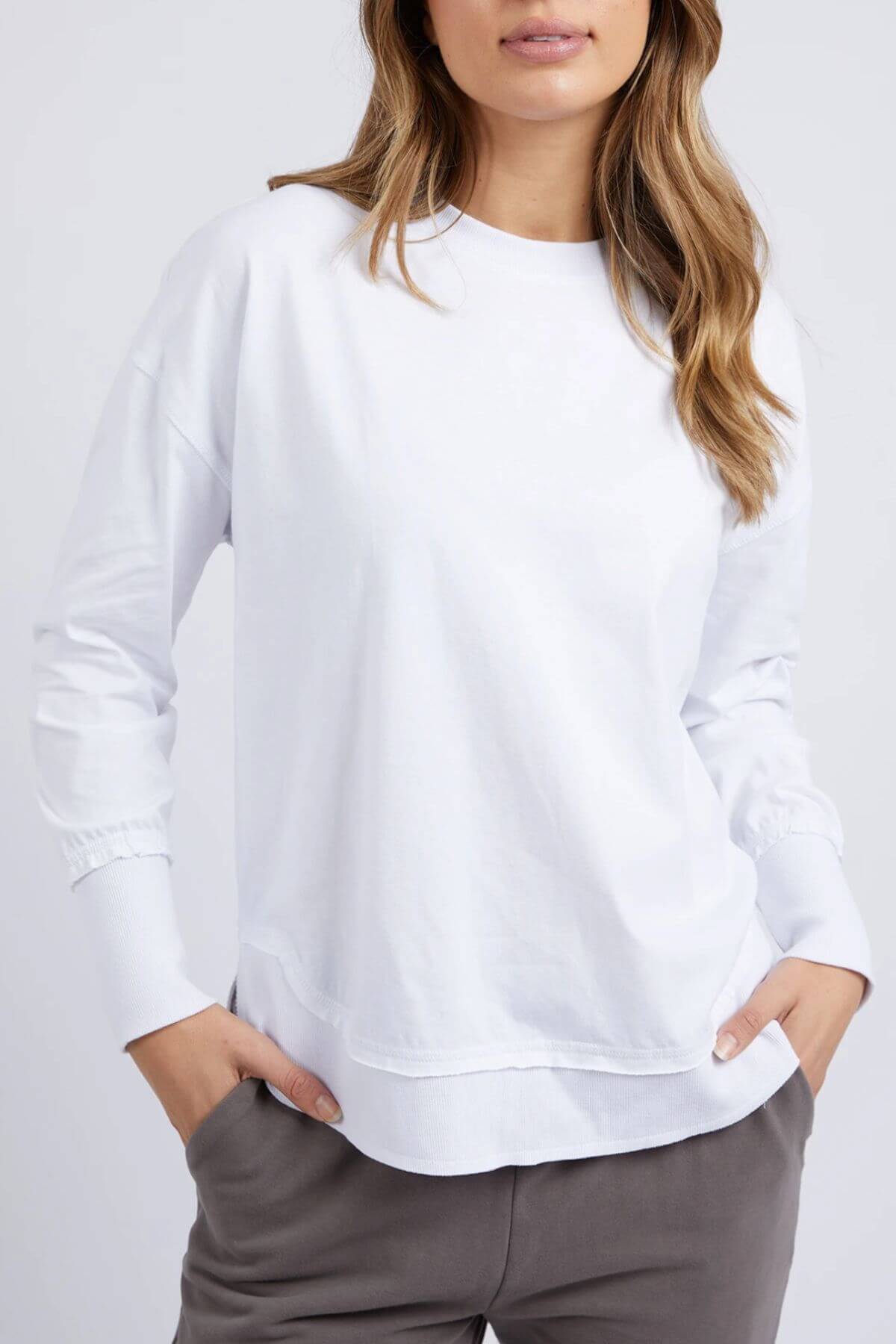 A woman models the Foxwood Farrah Long Sleeve top with gray pants, hands in her pockets. Only her upper body is visible, highlighting the shirt’s relaxed fit.