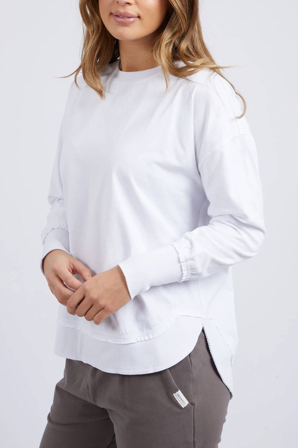 A woman with long, light brown hair wears the Foxwood Farrah Long Sleeve top and gray pants, standing against a neutral background with her hands loosely clasped.