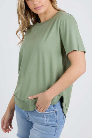 A person wearing a loose-fitting Foxwood Farrah Short Sleeve Tee, paired with light blue denim jeans. The individual has long, wavy hair and is shown from the waist up, with one hand placed in their pocket. The background is plain and light-colored.